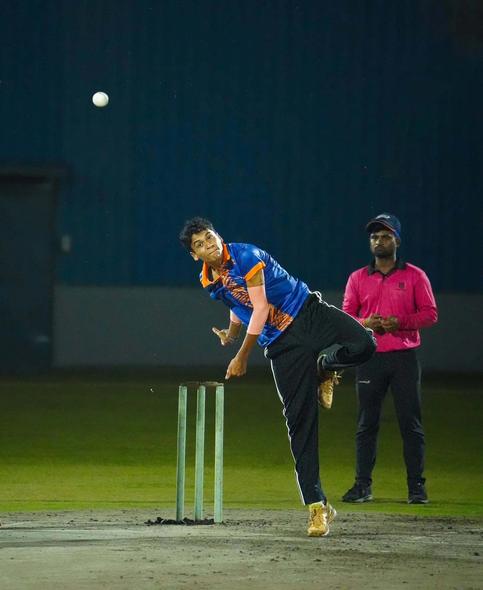 An SCA bowler in his follow-through under floodlights, the umpire behind him