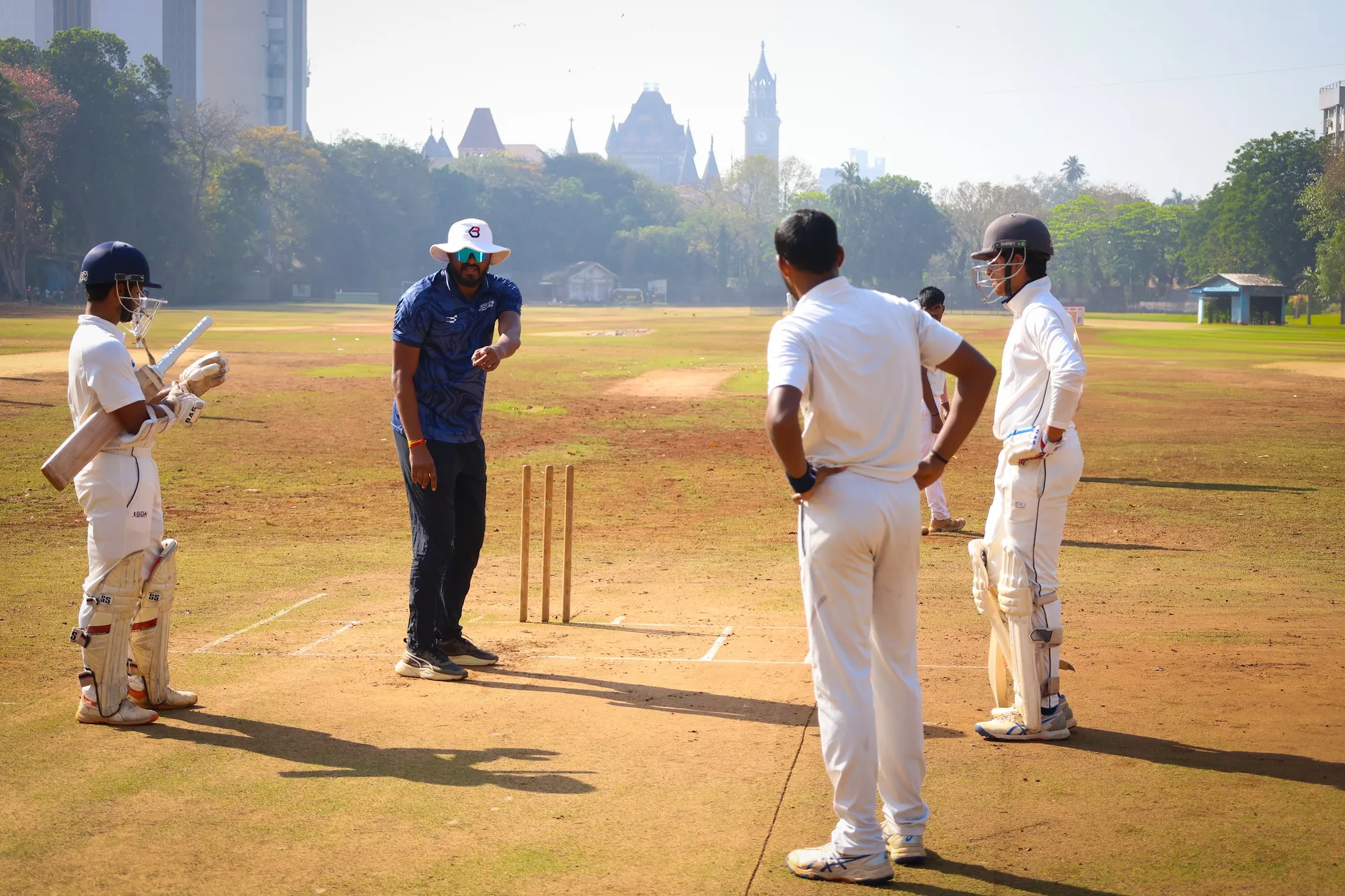 A coach talking with players in the middle of the pitch, the Mumbai skyline behind