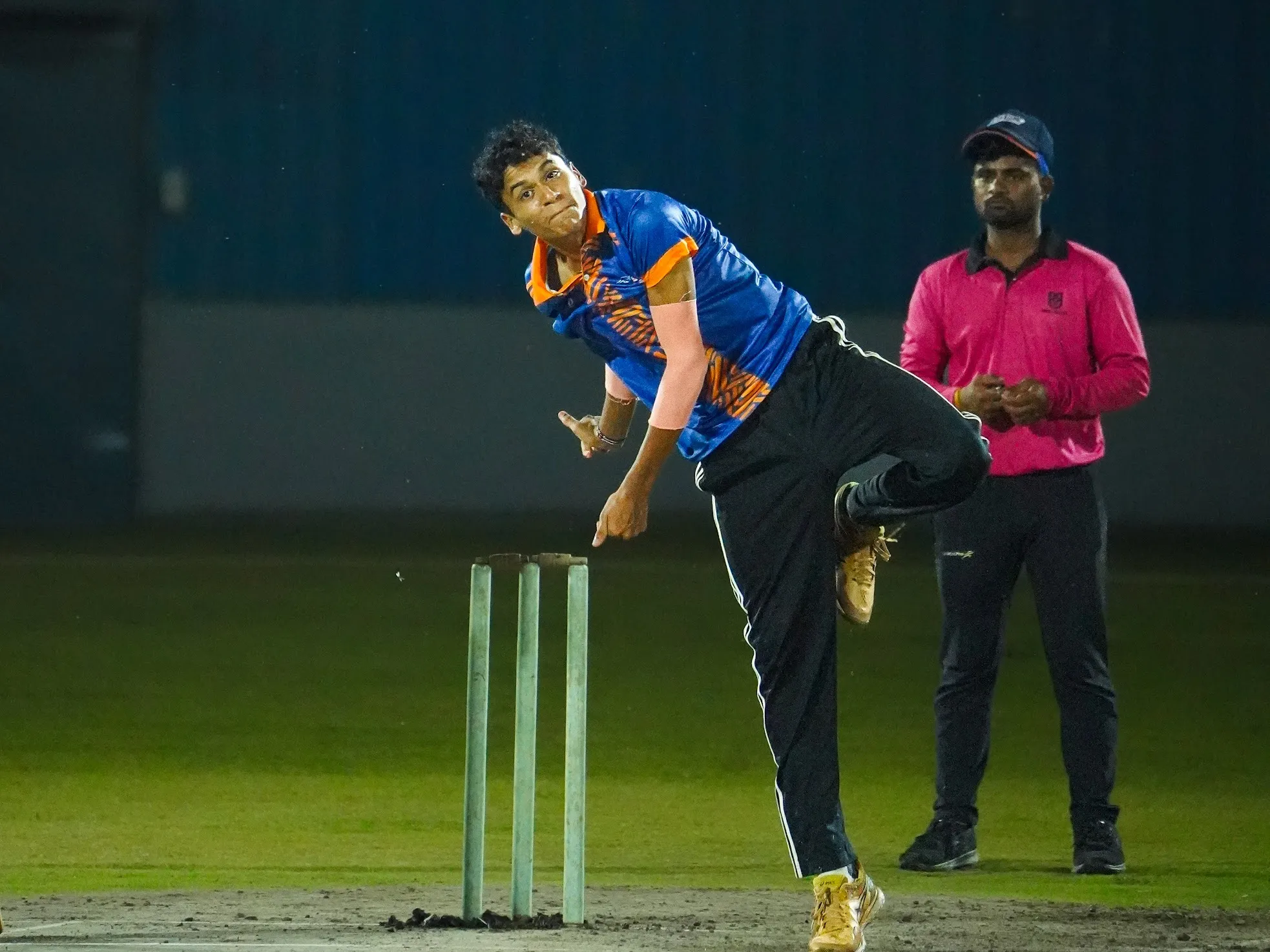 An SCA bowler in his follow-through under floodlights, the umpire behind him