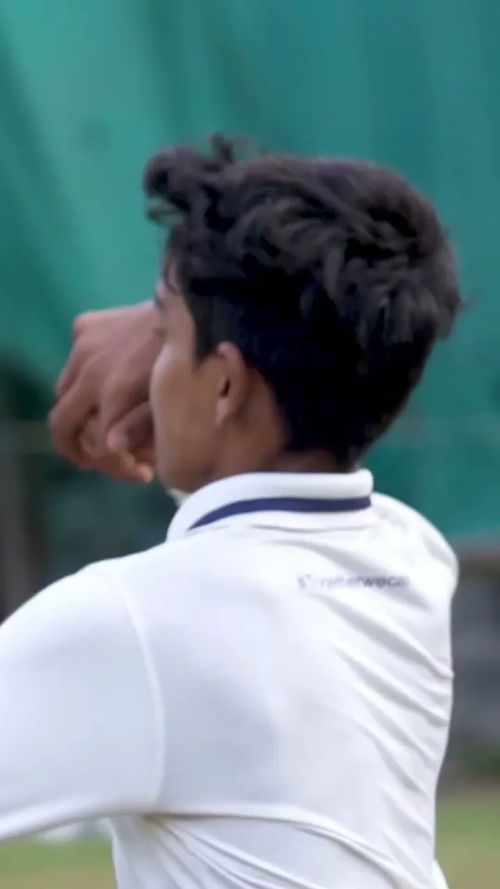 A young bowler in a white shirt mid-delivery in front of the nets.